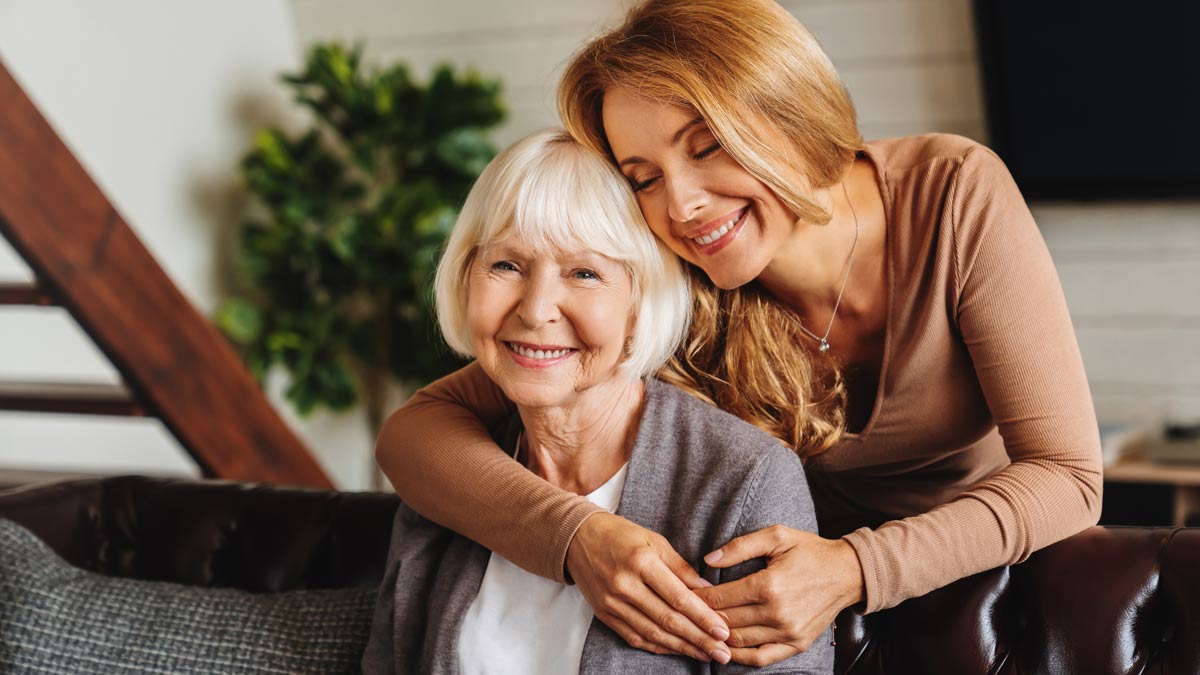 Happy daughter embracing from behind elderly mother at living room.